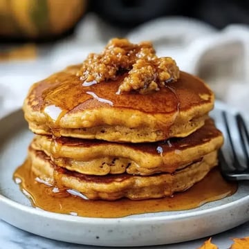 A stack of fluffy pancakes topped with syrup and a dollop of butter, served on a light-colored plate.